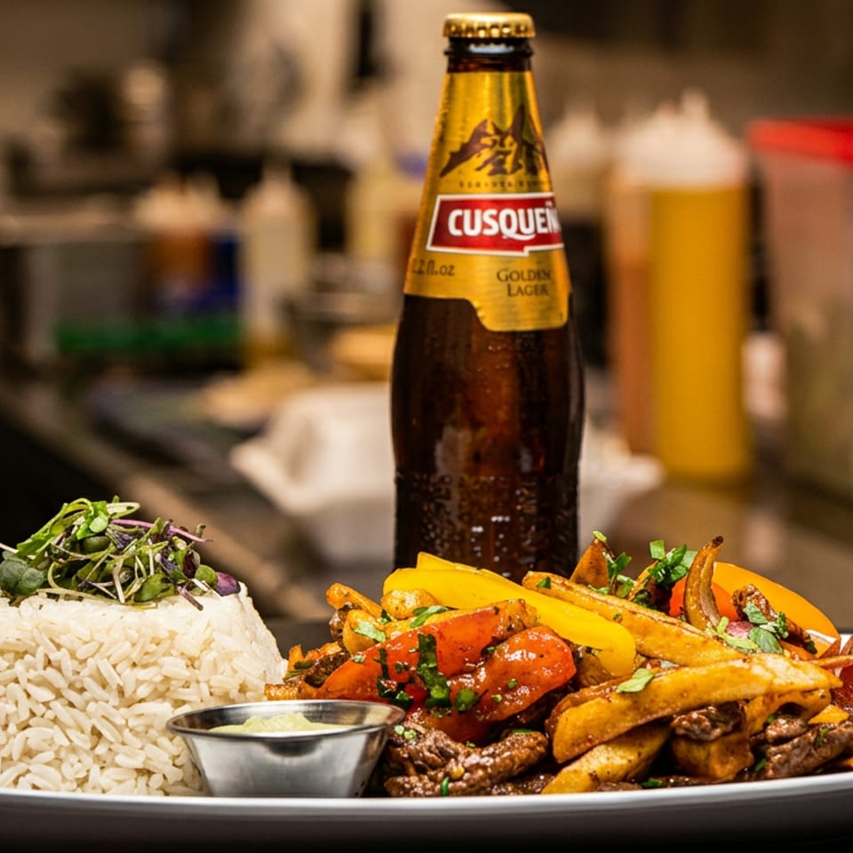 Plate of Lomo Saltado with rice, fries, and a bottle of Cusqueña beer in a restaurant kitchen.